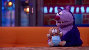 Een foto van pop Frits Fladder die op een bruine bank aan een bruine tafel zit. Hij is een vleermuis met twee scherpe tanden en rechtopstaande oren. Hij is paars op de rug en lichtpaars van voren. Hij heeft een bruine knuffelbeer met blauwe kleding vast.