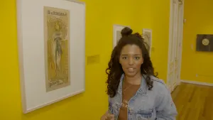 Een foto van presentator Sosha. Ze staat in een museum, met gele wanden, en kijkt in de camera. Naast haar, aan de muur, hangt een reclameposter in de kunststijl Art nouveau. Dat ziet er sierlijk uit.