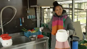Een foto van presentator Sosha. Ze staat in een keukentje van Stichting Aap. Ze houdt een plastic pot vast en kijkt in de camera. Op het aanrecht staat een kist met groente.