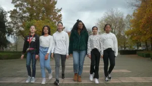 Een foto van presentator Sosha. Ze staat tussen vijf meisjes uit groep acht. Ze lopen allemaal op een zebrapad in de richting van de camera.