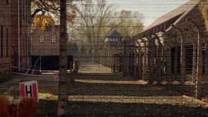 Een foto van concentratiekamp Auschwitz in Polen. Er is een hoog hek van prikkeldraad te zien vooraan, dat rechts doorloopt langs een gebouw met een schuin dak en een uitkijktoren verder naar achteren. Links zijn gevels van twee andere gebouwen te zien.