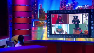 Een foto met twee poppen: Debbie van Dubben die zit, en een pop van een presentator die staat. Ze kijken met hun rug naar de camera naar een beeldscherm met daarop vier beelden van poppen: een brandweer, een politieagent, een dokter en een goochelaar.