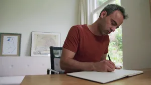 Een foto van de schrijver Gideon Samson. Hij zit aan een tafel en schrijft iets in een boek.