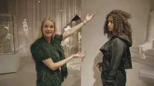 Een foto van presentator Janouk die in een kledingwinkel naast een pop staat en die laat zien door met haar handen ernaar te wijzen. De pop draagt een zwarte jas.