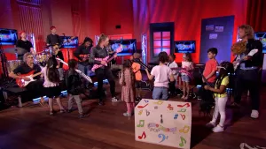 Een foto van presentator Pepijn in een Tv-studio. Hij staat samen met een vrouw tussen kinderen. Ze hebben allemaal een gitaar vast.  Links in beeld zie je een muziekband.