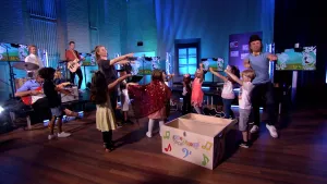 Een foto van presentator Pepijn in een Tv-studio. Hij heeft een hoed op en staat tussen kinderen. Ze zijn aan het dansen. Links in beeld zie je een muziekband.