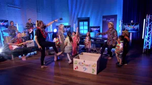 Een foto van presentator Pepijn in een Tv-studio. Hij heeft een hoed op en staat tussen kinderen. Ze zijn aan het dansen. Links in beeld zie je een muziekband.