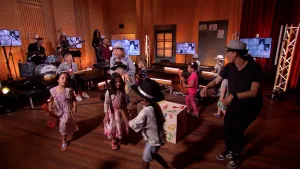 Een foto van presentator Pepijn in een Tv-studio. Hij heeft een hoed op en staat tussen kinderen. Ze zijn aan het dansen. Links in beeld zie je een muziekband.