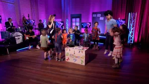 Een foto van presentator Pepijn in een Tv-studio. Hij heeft een pruik met zwarte krullen op en staat tussen een groep kinderen. Ze zijn aan het dansen. Links in beeld zie je een muziekband. 