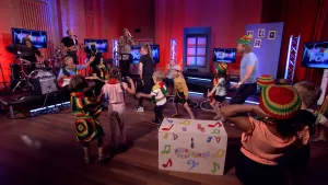 Een foto van de Apennoten Pop studio. Middenin de studio zijn kinderen aan het dansen en op de achtergrond is er een muziekband die aan het spelen is. 