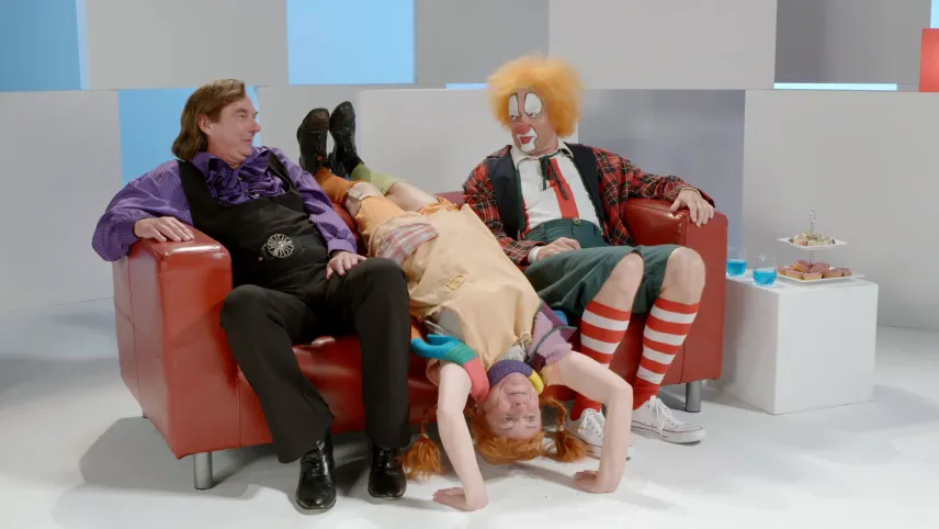 Een foto van drie acteurs in een studio. Ze zitten op een bankstel. Twee van hen spelen Bassie en Adriaan. De derde speelt Pippi Langkous. Pippi hangt ondersteboven op het bankstel.