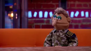  Een foto van pop commandant Winters op een bruine bank achter een bruine tafel. De pop heeft een blouse aan met een legergroen en grijs patroon en een zwart met geel gestreept stuk stof op de schouders. De pop heeft een lange, dunne neus en bruin haar.