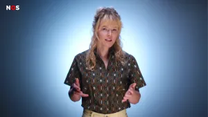  Een foto van een presentator die een lichtbruine broek aanheeft en een zwarte blouse draagt met bruine lijntjes erop waarin steeds vier groene blaadjes staan met plusteken ertussen. De achtergrond is blauw.