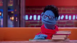 Een foto van pop Boeke Legger op een bruine bank met voor zich op een bruine tafel een stapel van vijf boeken en één boek waar de handen van de pop opliggen. De pop heeft een blauwe huid en zwart haar. De pop draagt een rode trui en een paarse bril.