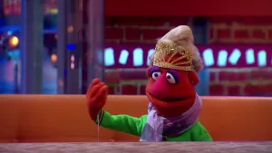 Een foto van pop Prinses Minima, waarvan de hand een zwaaiende beweging maakt. De pop heeft een rode huid, lichtblond haar en paarse oogschaduw op. De pop draagt een groene trui, een paarse sjaal en om haar hoofd een goudkleurig kroontje. 