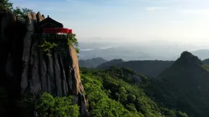 Een foto genomen in Zuid-Korea van een bruine, stenen berg met van veraf een houten huisje met een schuin dak waar een soort palen met rode bollen omheen staan. Onderaan de berg en op bergtoppen en heuvels erachter, staan groengebladerde bomen en planten.