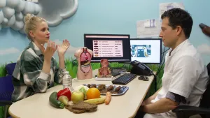 Een foto van Janouk en Tim met op tafel: twee beeldschermen met menselijke poep en poep binnenin de darmen erop, een dienblad met poep voorbeelden, een plastic menselijk lichaam met organen,  groenten, fruit, een fles water en twee sneetjes volkorenbrood.