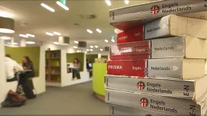 Een foto van een ruimte in een school. Op de voorgrond zie je een stapel woordenboeken, van Engels naar Nederlands. Op de achtergrond zie je mensen zitten.