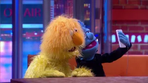 Een foto van pop Anton Konijn die een selfie maakt met zijn witte mobiele telefoon samen met de pop van de presentator. Anton konijn heeft een blauwe huid, een zwart pak en een stukje van zijn blauwe haar is te zien. De presentator heeft een gele vacht.