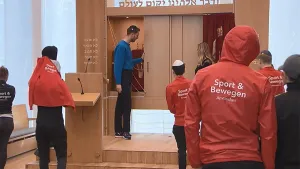 Een foto van mensen in een synagoge. Achterop hun jasjes staat (bij sommige) de tekst 'Sport & Bewegen Amsterdam'. Enkele van de mensen hebben een keppel op. 