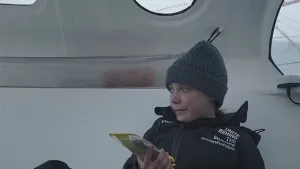 Een foto van Greta Thunberg, met een muts op. Ze zit in een ruimte met witte wanden en ronde ramen. Daarachter zie je sneeuw. 