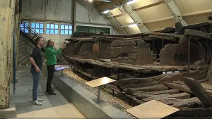 Een foto van twee mannen. Ze staan in een loods. Daarin zijn de restanten van een hele oude deels vergane boot tentoongesteld. 