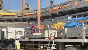 Een foto van een stadion in aanbouw. Je ziet een vrachtwagen met betonplaten en enkele bouwvakkers. Ze staan in het stadion dat gebouwd wordt.