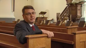 Een foto van een man die op een bank zit in een kerk. Op de achtergrond zijn er meer banken te zien en daarachter het podium.