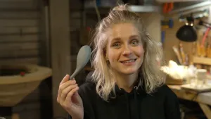 Een foto van presentator Janouk. Ze staat in een ruimte met allemaal gereedschappen en toont een kunststof lepel aan de kijker. 