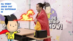 Een animatie van striptekenaar Leon die achter een bureau staat. Hij heeft een soort rode cape om. Links in beeld zie je zijn stripfiguur die zegt: 'Hij denkt zeker dat hij superman is'. 