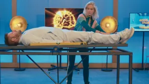 Een foto van presentator Job die op een tafel ligt die kan wankelen. Presentator Janouk staat achter de tafel. Op de tafel ligt een waterpas. Een waterpas is een meetinstrument met water en lucht om te zien hoe recht iets is.