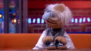 Een foto van pop Professor Bolleboos. De pop heeft wit haar, een gerimpelde lichtbruine huid op zijn gezicht, een bril op en een blauwe neus. De professor heeft twee blauwe sportschoenen met witte veters vast die voor hem staan op een bruine tafel.