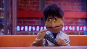 Een foto van pop Klaas Gadget die een afstandsbediening vastheeft zoals van een televisie. Hij heeft zwart, krullend haar en een gele bril op. Zijn blouse heeft blauwe en witte strepen. Hij zit op een bruine bank achter een bruine tafel.