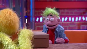 Een foto van pop Ab van Helsen in een blauwe blouse met een rood t-shirt eronder. Ab heeft lichtgroen haar en wijd opengesperde ogen met kleine pupil. Een kartonnen doos voor de pop op tafel wordt opengemaakt door een pop met gele vacht links in beeld.