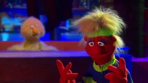 Een foto van pop Helga Commandant in een paars met groen trainingspak en een groene zweetband om het hoofd. De pop heeft een rode huid, omhoogstekend, lichtgekleurd haar en een ronde neus. Achteraan is onscherp een pop met gele vacht te zien.