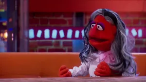 Een foto van een pop die niet bij naam genoemd wordt, maar "een moderne heks". Ze heeft een rode huid, haar dat deels zwart en deels grijs is. Ze draagt een witte blouse en een rode strik om haar nek. Ze zit op een bruine bank achter een bruine tafel.