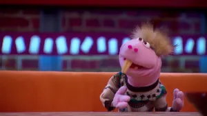 Een foto van pop Walt van het Slob op een bruine bank achter een bruine tafel. De pop heeft een roze huid, rechtsopstaand lichtbruin haar, twee lange, gele voortanden zoals een bever en een witte trui met groene en rode vormen erop aan.