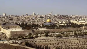 Een foto van de stad Jeruzalem met in het midden de in de islam heilige Al-Aqsamoskee, een grijs gebouw met goudkleurige koepel erop en groene bomen ervoor. Daarvoor staat een lichtbruine muur, de buitenkant van de, in het jodendom heilige, klaagmuur.
