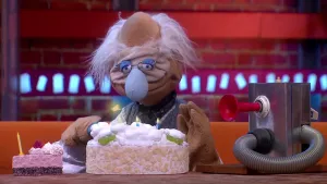 Een foto van pop professor Bolleboos met grijs haar, een rimpelig gezicht en een lichtblauwe neus. Voor hem op tafel staan een kleine, roze taart, een witte taart met brandende kaarsjes en een machine met een slang eraan en een rode toeter erop.