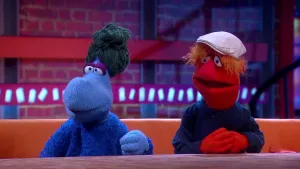 Een foto van poppen Tinus en Trees Veldhoen op een bruine bank achter een bruine tafel. Trees heeft een lichtblauwe huid, donkergroen haar en een donkerblauwe trui aan. Tinus heeft een rode huid, blauwe trui aan, oranje haar en een witte pet op.