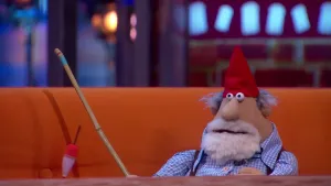 Een foto van pop Klaas Wouter Spillebeen op een bruine bank achter een bruine tafel. De pop heeft een rode puntmuts op, grijs haar, een witte baard, een blauw met wit geblokte blouse aan en een vishengel van bamboe met rood met witte dopper vast. 