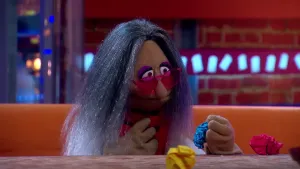 Een foto van pop Emmy Kwast op een bruine bank achter een bruine tafel. De pop heeft dun, grijs haar, een roze zonnebril op en een rode sjaal om. De pop een blauwe prop papier vast. Op tafel ligt ook een rode prop papier en een gele prop papier.