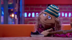 Een foto van pop Sil de strandjutter op een bruine bank achter een bruine tafel. Sil heeft een blauw en lichtbruin gestreepte muts op en een bruin stuurwiel van een schip vast, dat op een roze zwembroek ligt. Een paarse sportschoen staat ernaast.   