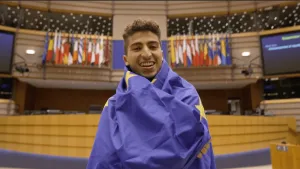 Een foto van presentator Nizar die de Europese vlag om zijn lichaam heeft terwijl hij in de kamer staat van het Europese bestuur. Op de achtergrond zijn er vlaggen zichtbaar van landen in de Europese Unie.