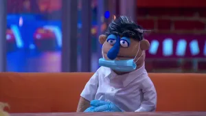 Een foto van pop tandarts Edo Bruins in een witte blouse en met lichtblauwe plastic handschoenen. Een lichtblauw mondmasker hangt onder zijn mond, vastgemaakt met witte touwtjes om zijn oren. Zijn huid is bruin, neus en ogen blauw en haar zwart met grijs.