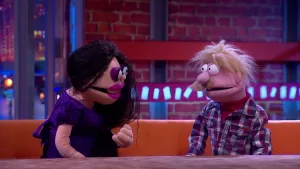 Een foto van poppen Annechien en Rob op een bruine bank achter een bruine tafel. Annechien heeft zwart haar, een paarse jurk, grote lippen met paarse lippenstift en paarse oogschaduw. Rob heeft blond haar, een snor en een paars en wit geblokte blouse.