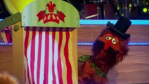 Een foto van pop signor Olifante die een rode huid heeft, bruin hoofd- en baardhaar en een zwarte snor. De pop draagt een zwarte hoge hoed en een groene sjaal met stippen. Naast de pop staat een soort poppenkast met een rood en wit gestreept doek ervoor.