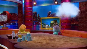 Een foto van een pop met gele vacht en pop meneer Fris die een lichtblauwe vacht heeft en paars haar. Meneer Fris heeft een rood touw vast met een witte, in de lucht zwevende, wolk eraan. Ze zitten op een bruine bank aan een als boterham gevormde tafel.