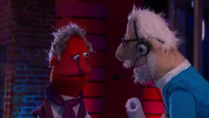 Een foto van pop roddelkoning Robert Achterklap met een rode huid, grijs haar en paarse sjaal en jasje. Ernaast een pop met wit haar, een blauwe trui en een zwarte bril die een hoofdtelefoon met microfoon opheeft en opgerolde witte papieren vastheeft.