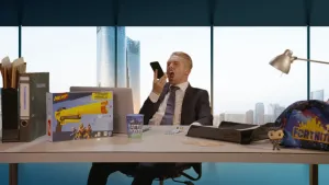 Een foto van een man achter een bureau op een kantoor. Hij houdt zijn mobiel in een hand en schreeuwt ertegen. Op zijn bureau staat speelgoed. Op de achtergrond zie je een moderne wolkenkrabber.